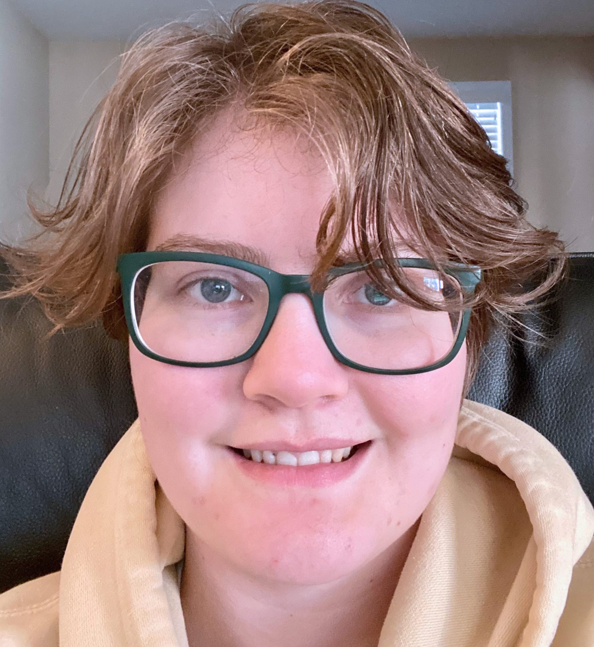 Young college student with short wispy reddish brown hair. They are wearing a light yellow hoodie, and has dark glasses. They are smiling. They appear to be sitting in a recliner or similar chair.