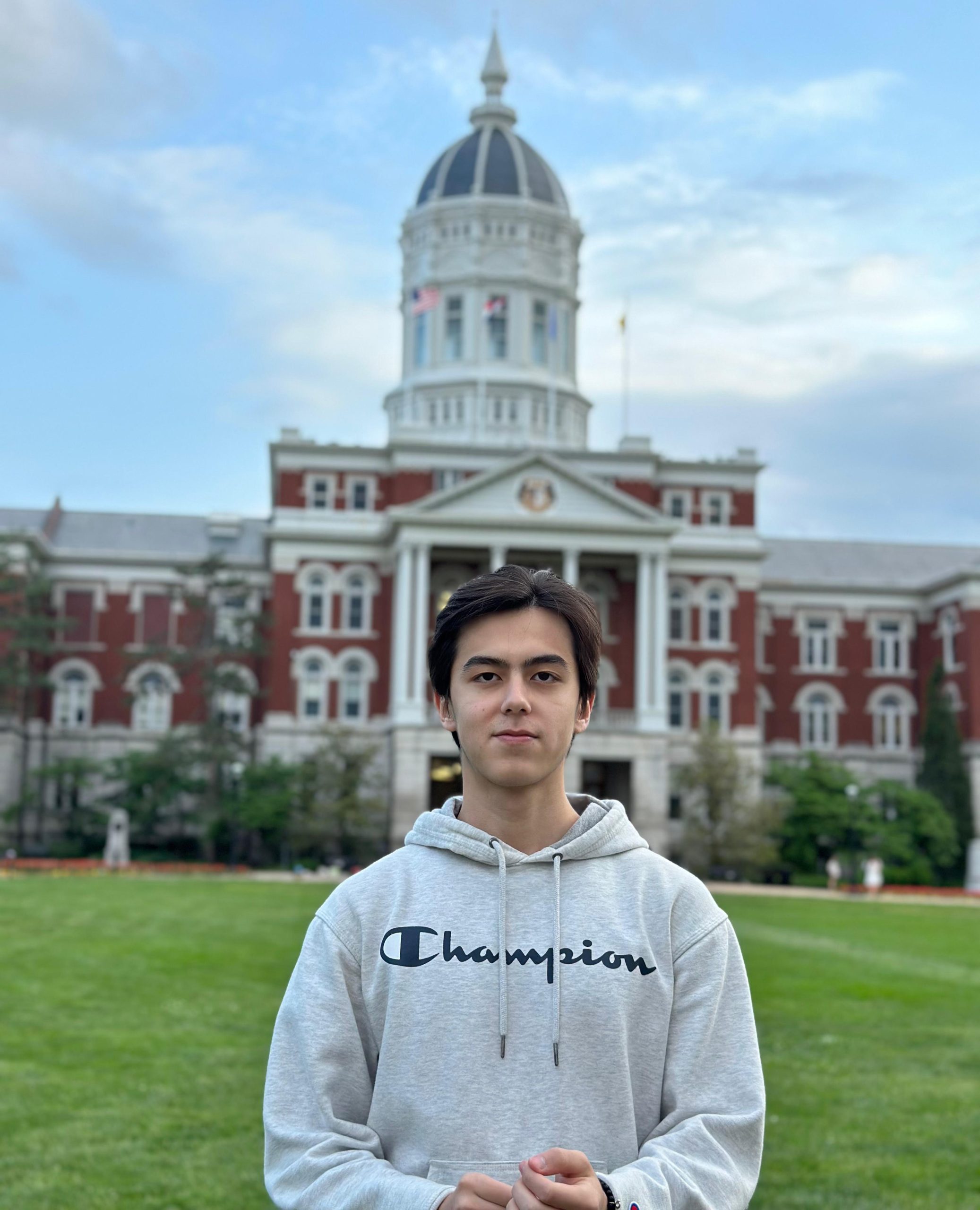 Male college student wearing a gray Champion hoodie standing in the grass in front of Mizzou's Jesse Hall.