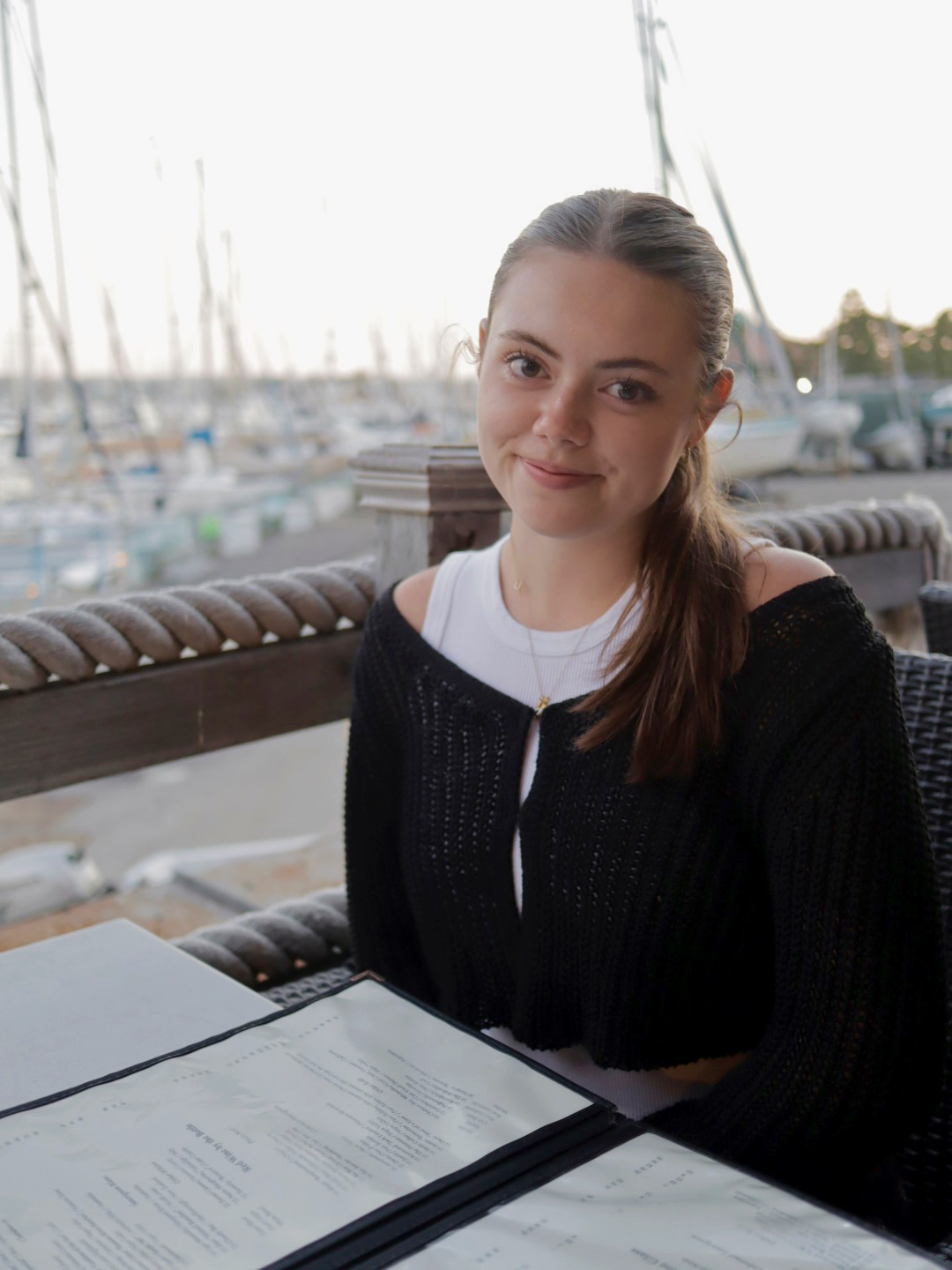 A young lady sitting at a table in front of the water and boats, with a menu in front of her.