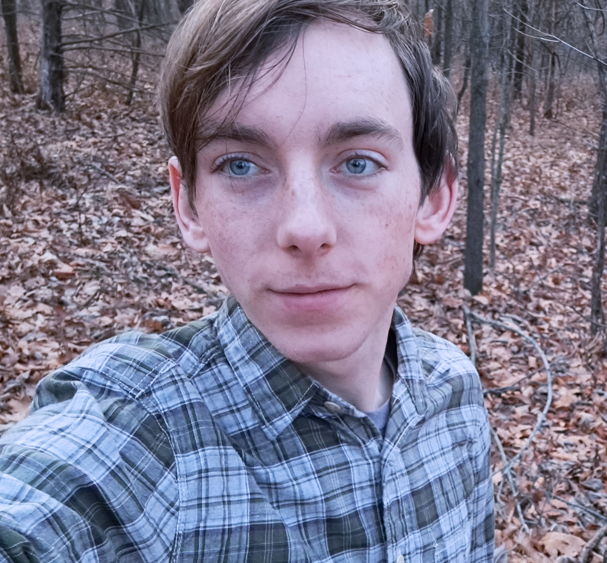 Young man in the woods during fall or winter with bare trees behind. He is taking a selfie, and is wearing a blue plaid shirt. He has light brown hair, parted on the side and blue eyes.