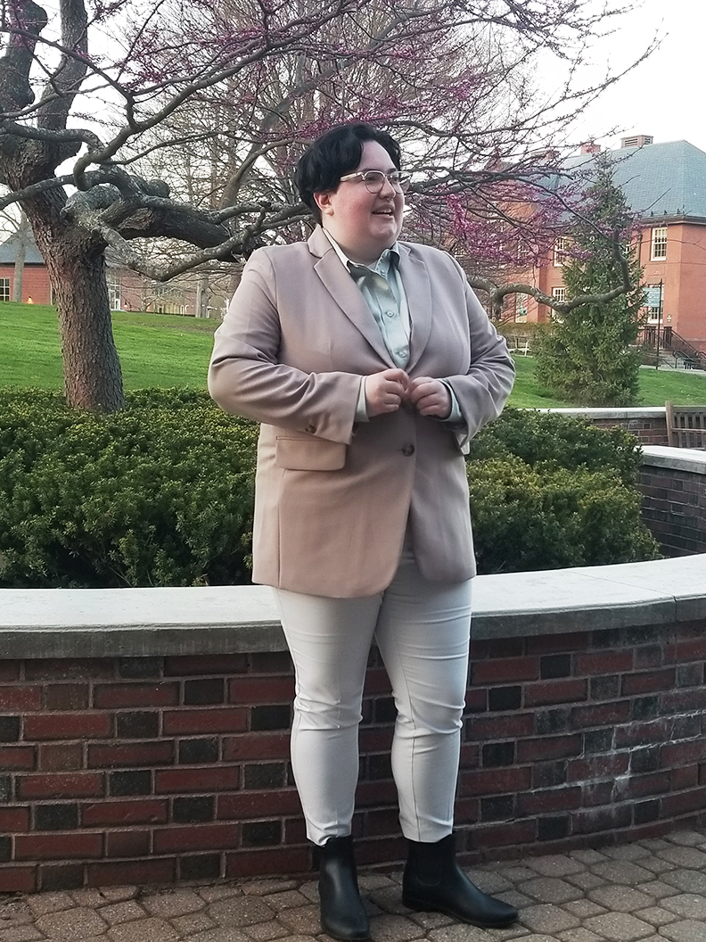 Person in a light pink suit jacket and white leggings standing in front of a short brick wall and bushes on campus.