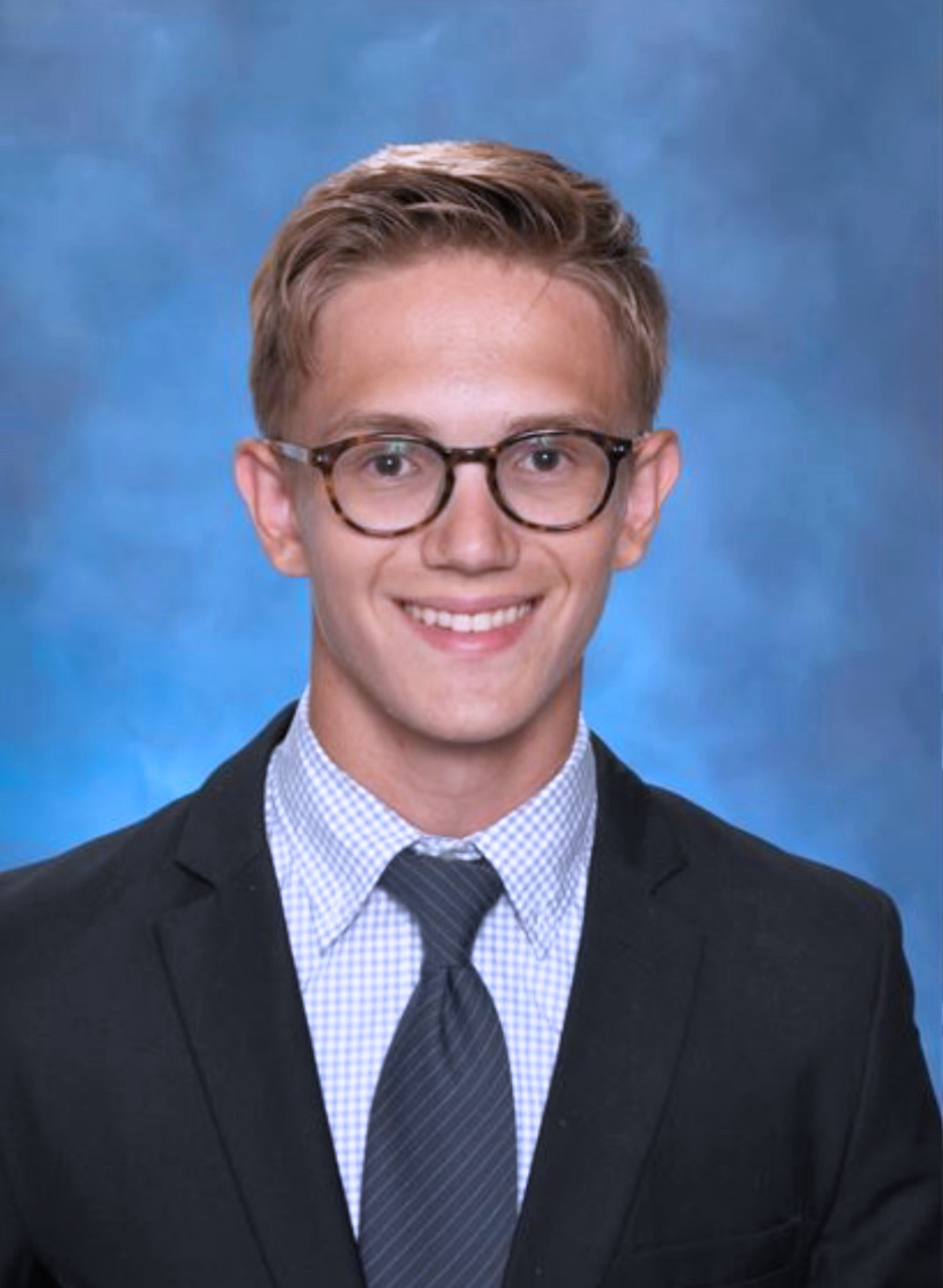 Young man with dirty blonde hair, wearing dark rimmed glasses and a black suit, with a dark blue tie and light blue pinstriped shirt. He is smiling. There is a blue background.