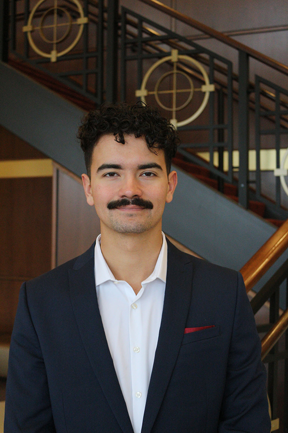 A college student wearing a suit jacket over a button-up white shirt. He has dark hair and a dark mustache. He is standing in front of a staircase.