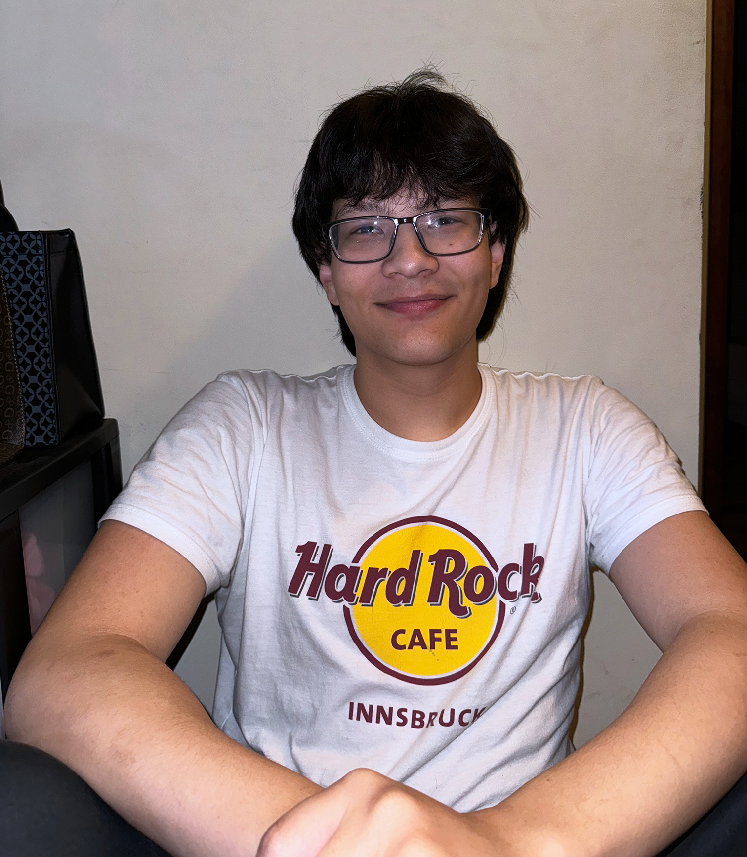 A male college student wearing a white Hard Rock Cafe - Innsbruck t-shirt. He has dark hair and glasses, and is grinning. He has brown skin. He is sitting in front of a white wall and has his hands held in front of him.