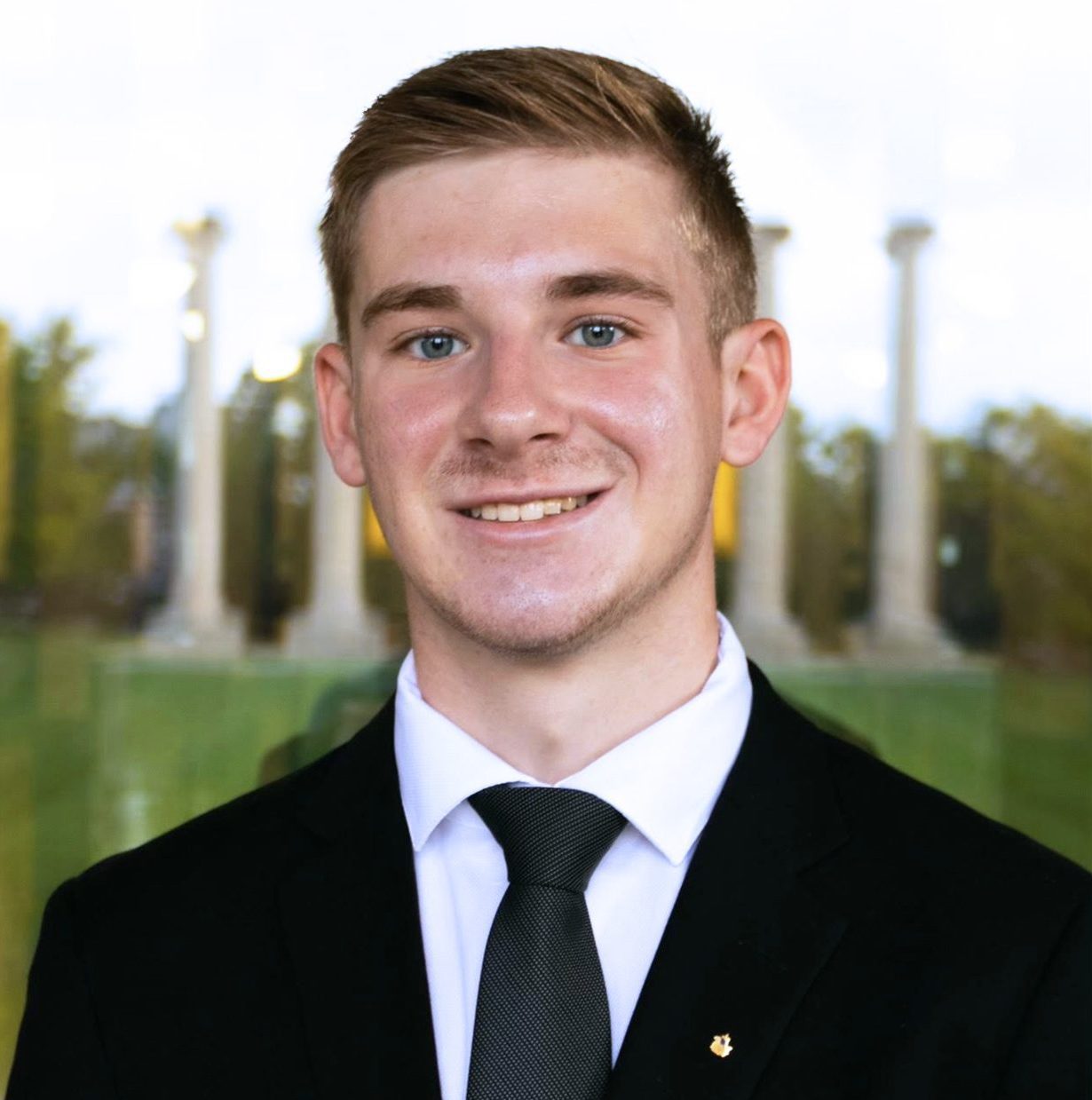 Professional headshot of a college student wearing a black suit with a white collared shirt and a navy blue tie. He is smiling and has light brown hair. He is standing before a background with the Jesse Hall columns of Mizzou.