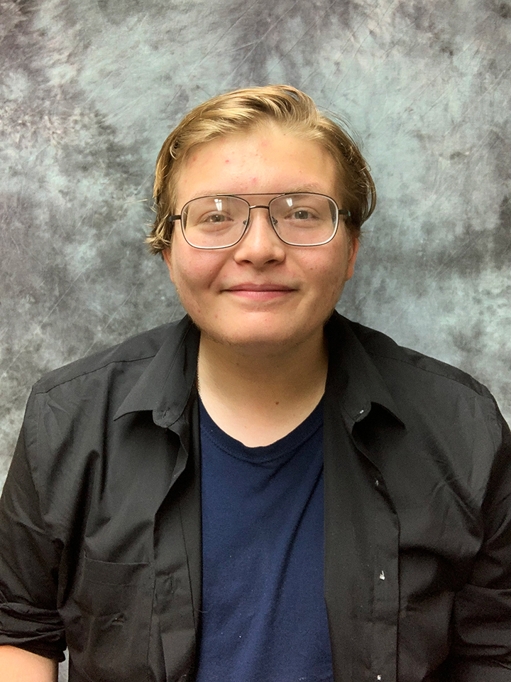 Young college student in front of a speckled photo background. Wearing a dark blue t-shirt with an open button-up black shirt over it. He is wearing glasses and has blonde hair. He is slightly smiling.