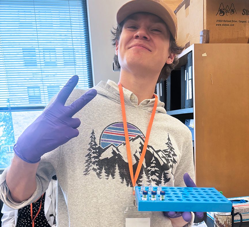 Male college student standing in a lab holding a plastic tray of test tubes. He is holding a peace sign with his other hand, on which he is wearing a latex glove. He is wearing a cap and a sweatshirt, along with an orange lanyard and name tag which is turned backwards. The young man is smiling.