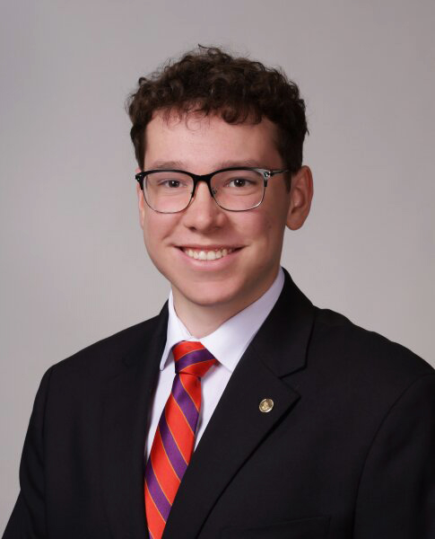 Young man in a black suit jacket and wearing a red and purple striped tie. He has brown hair and glasses that are dark-rimmed at the top. He is smiling. It is a professional photo.