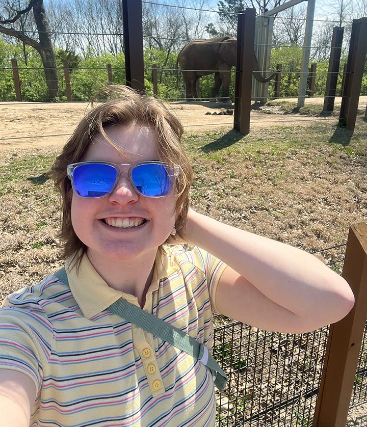 Young lady with short brown hair wearing reflective blue sunglasses. She has her hand behind her head and elbow bent. She is wearing a striped yellow polo shirt and has a purse strap across her chest. She is surrounded by leaves and bare trees with a bright blue sky.