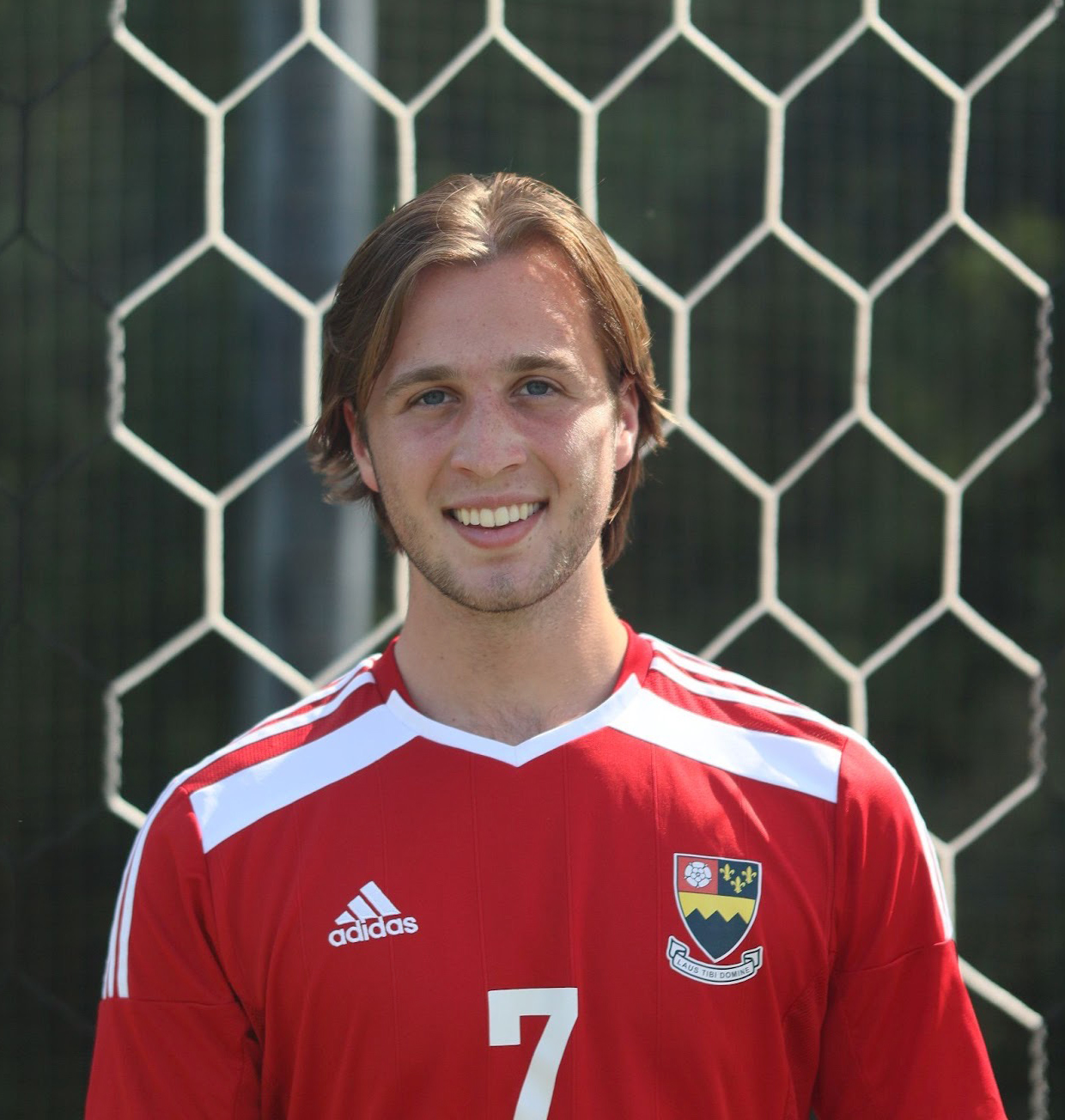 College soccer player in front of a net. He is wearing a red shirt with white accents and number. He has blonde hair and blue eyes.