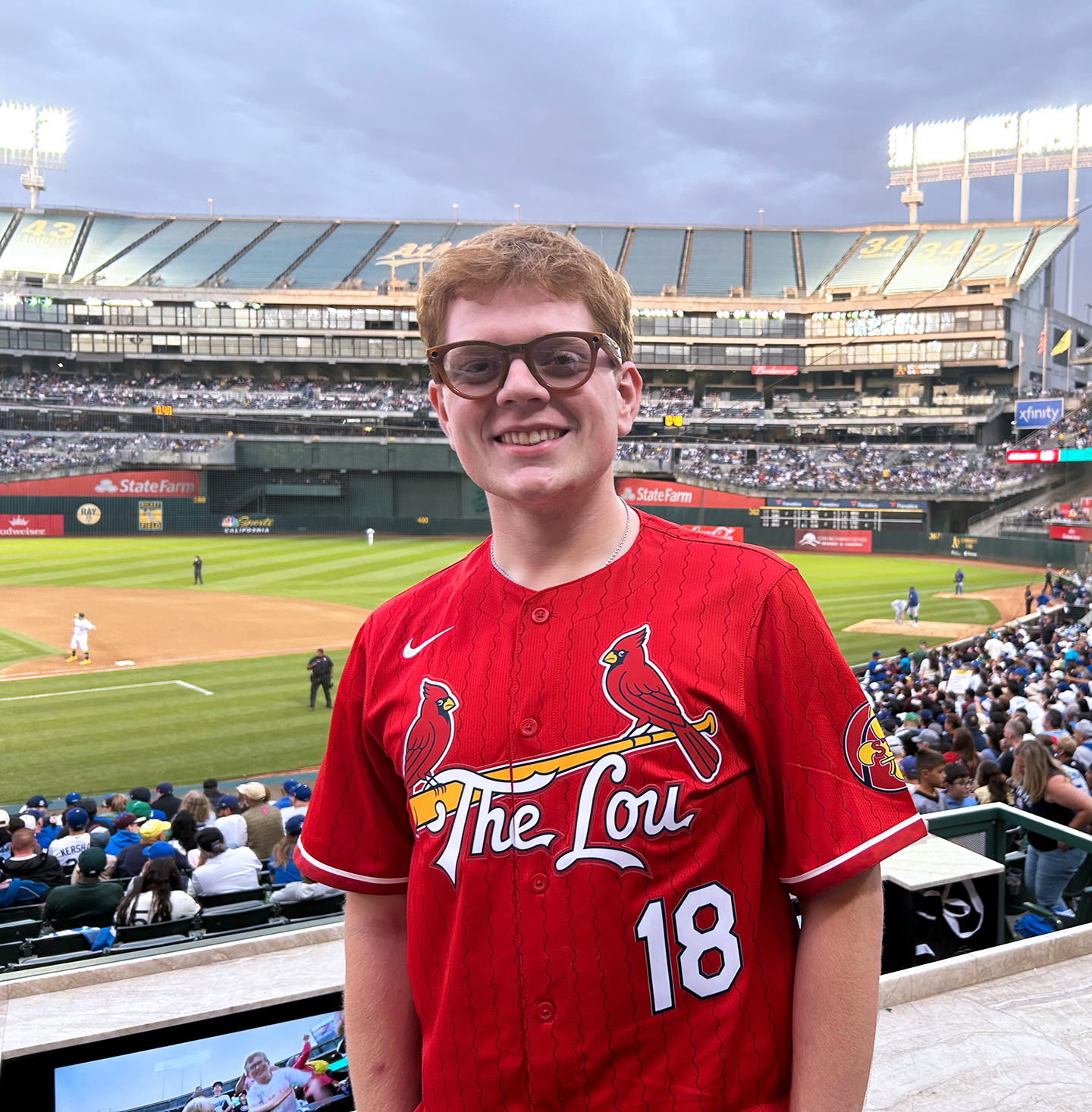 Male college student standing in the stands at St. Louis Cardinals baseball game, with the baseball diamond behind him. He is wearing a red jersey with 