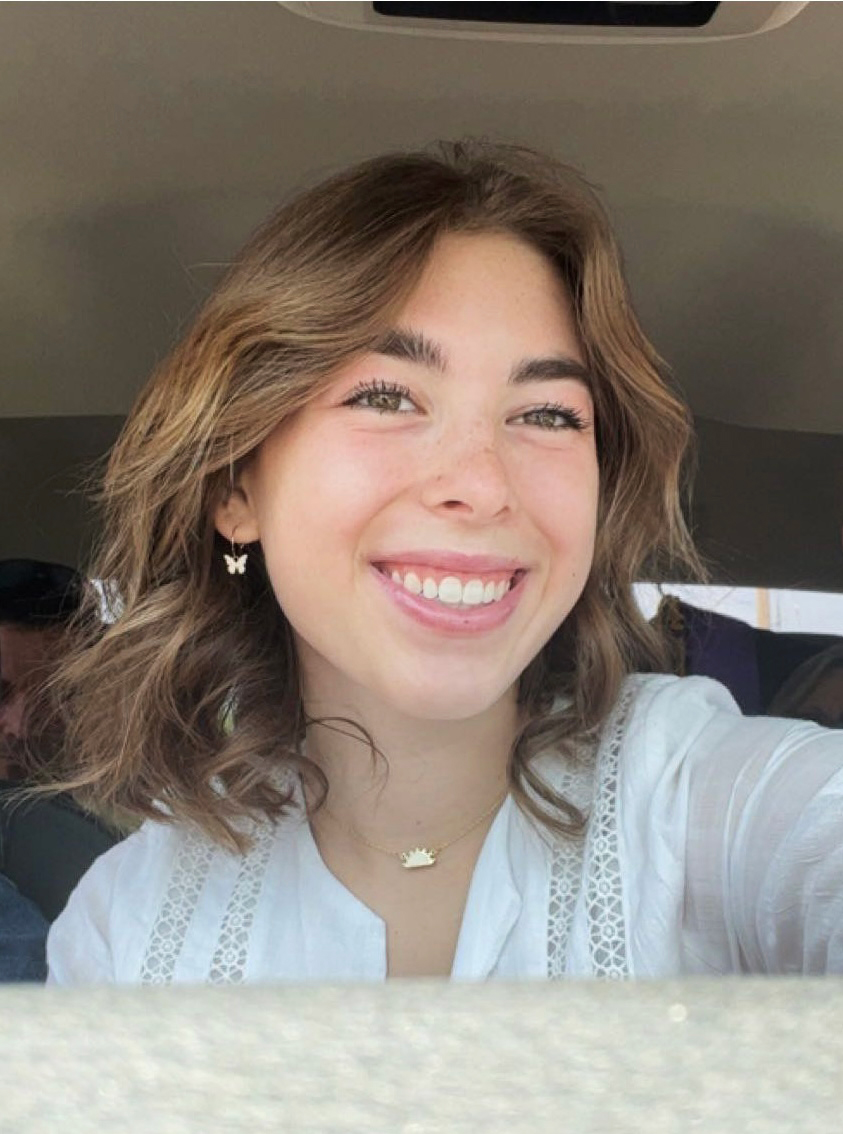 Young Asian lady smiling. She has medium brown hair, is wearing a white shirt, open at the collar with a necklace and earrings. She is in a vehicle with people behind her.