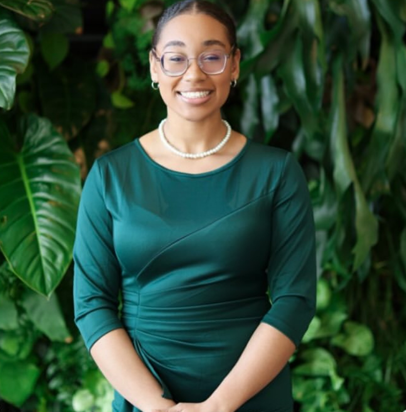 African-American female college student smiling in front of green plants while wearing a green satin top or dress.