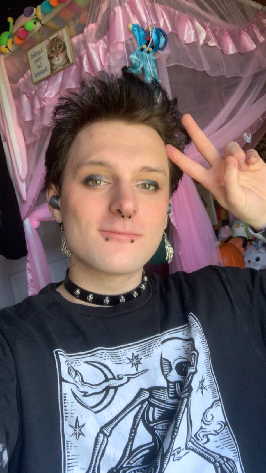 Alternative-looking college student giving a peace sign. He has a spiked black collar on and a black t-shirt. His bottom lip is pierced twice and he is wearing eyeliner. He has dark hair and ear piercings.