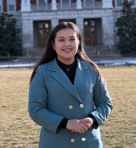 Female college student wearing a gray button up coat, standing in the grass in front of Mizzou's Jesse Hall. She has long dark straight hair and has her hands grasped in front of her. She is smiling.