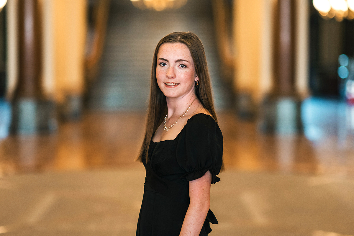 Thin young lady with long brown hair turned sideways facing the camera. She is wearing a black feminine shirt. The background is blurred but looks like a stage or staging area.