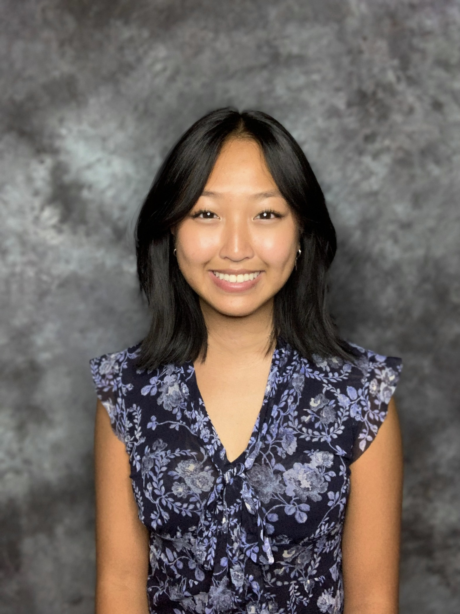 Asian college student with gray background who is wearing a blue floral shirt on a black background, earrings, and she is smiling.