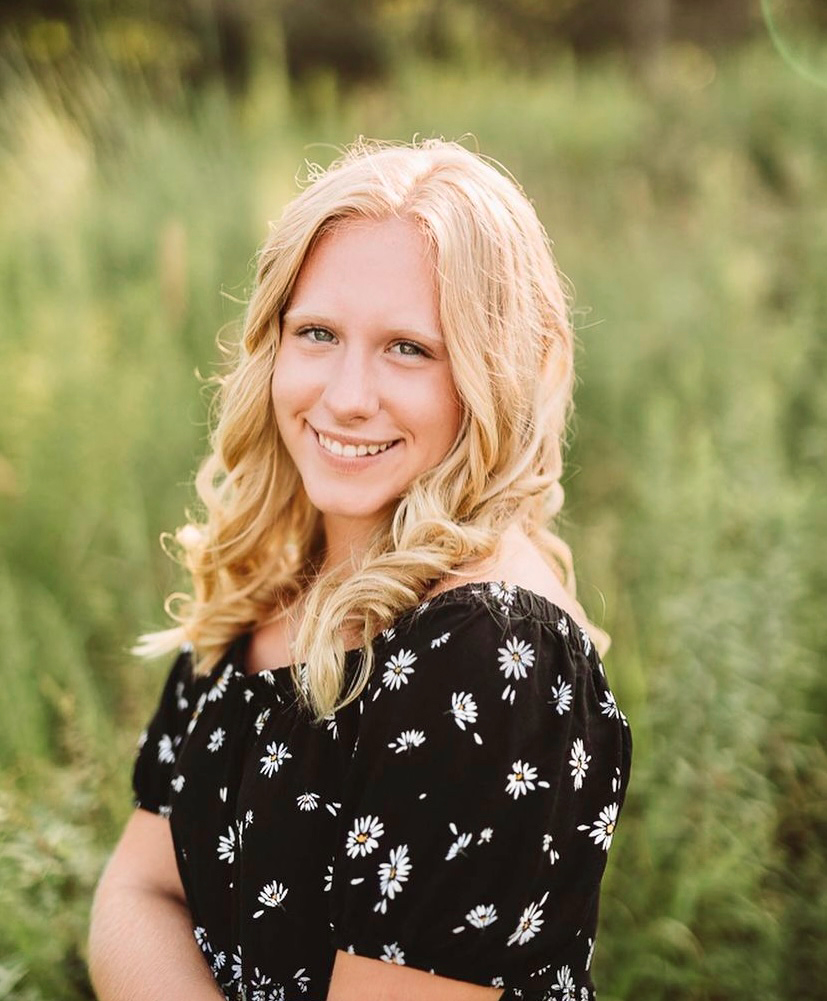 Young college girl with medium curly blonde hair smiling. She is wearing a black dress top with short fluttered sleeves and tiny white flowers. She has her arms crossed in front of her. She is posing in front of a blurred field.