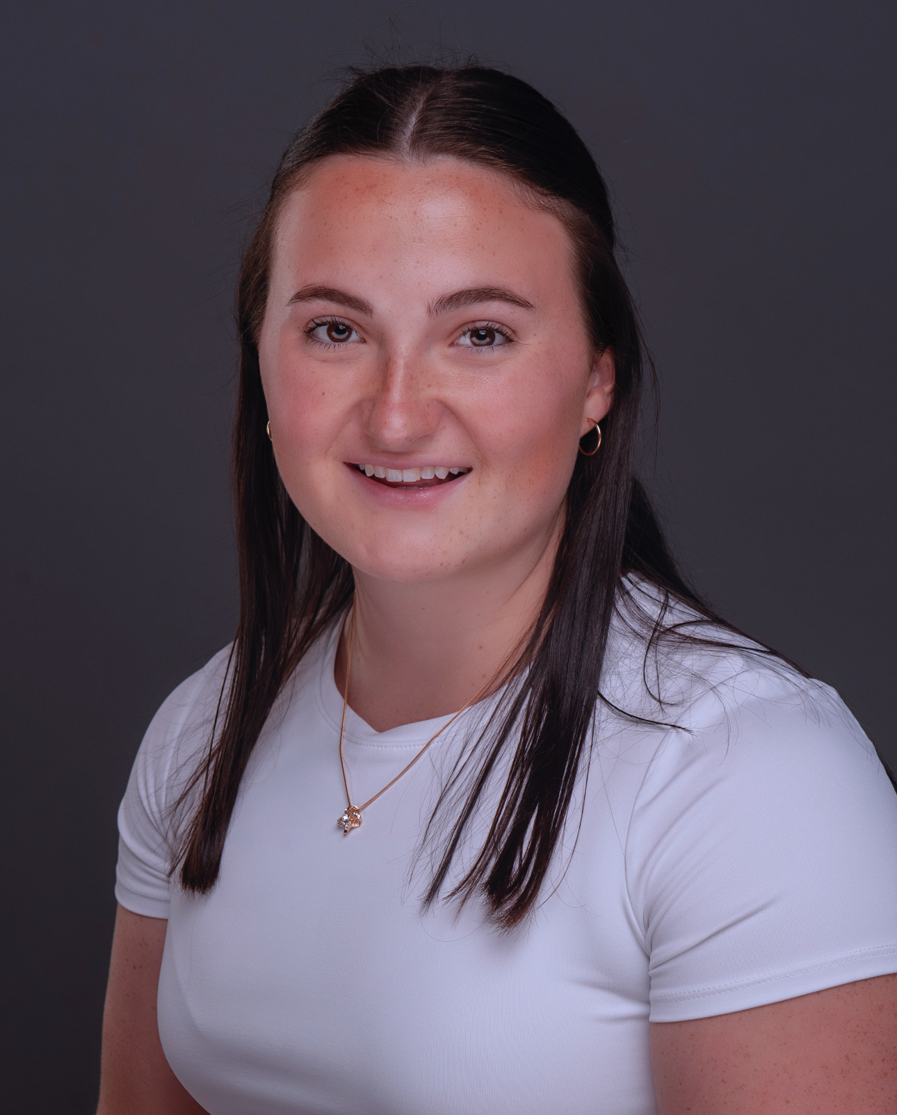 Young college girl with medium-long brown hair smiling. She is wearing a white tshirt and gold necklace.