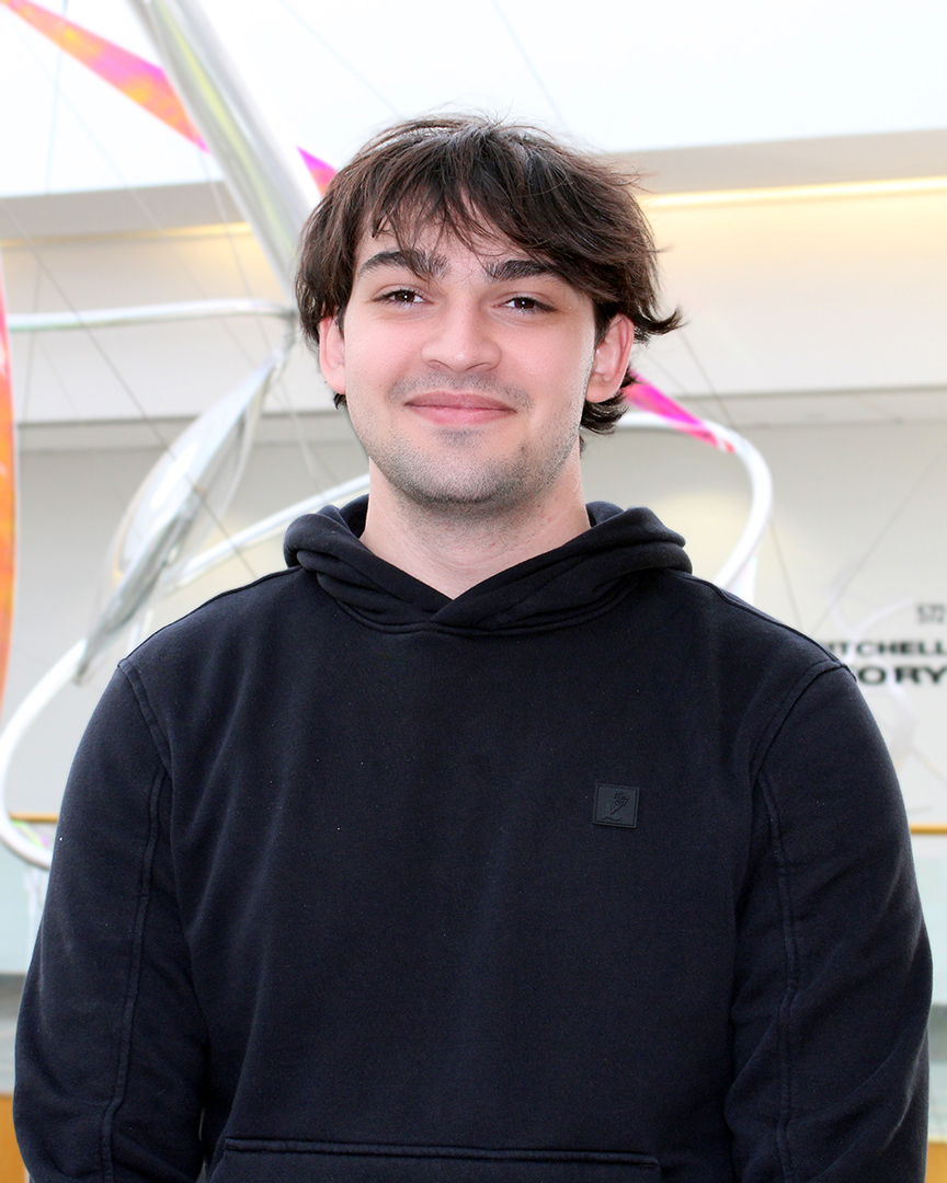 Young man standing smiling. He has dark hair and eyes and a bit of peach fuzz on his chin. He is wearing a black sweatshirt.