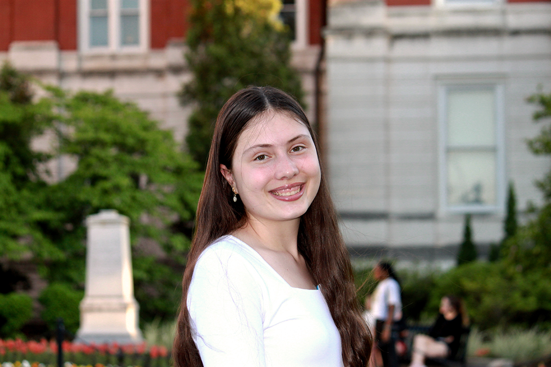 College Student standing in front of Jesse Hall. She has long dark hair and a white shirt and she is smiling.