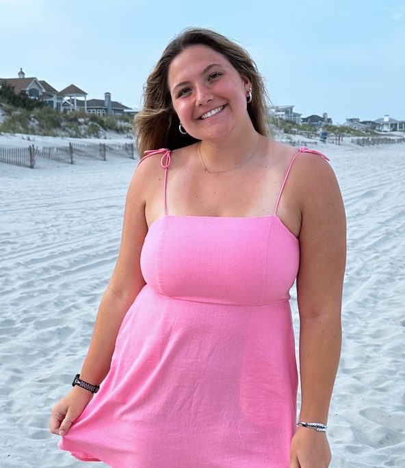 Young college student standing on a beach in a short pink sundress. She is smiling and grasping the bottom of her dress as if to hold it down. She has brown hair and has her head tilted to one side.