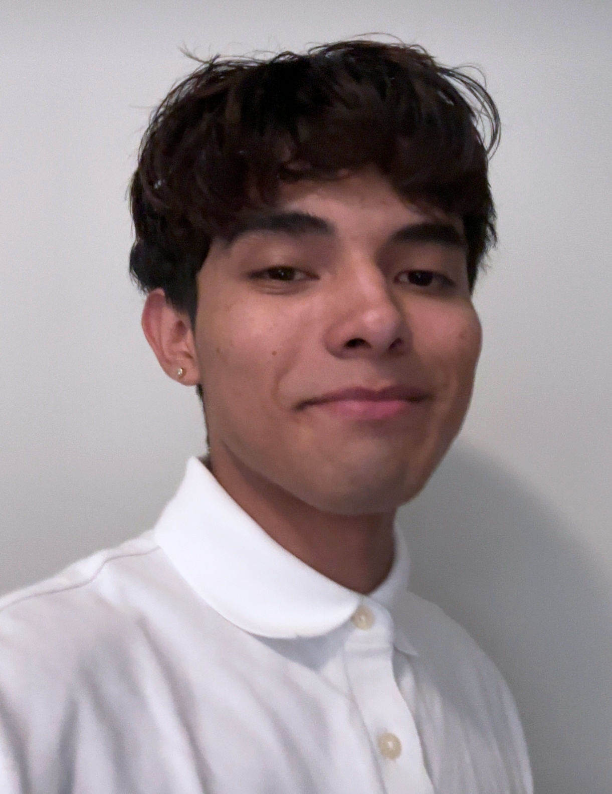 A male college student wearing a white polo shirt, buttoned. He has dark hair and is smirking. He has brown skin. He is standing in front of a white wall.