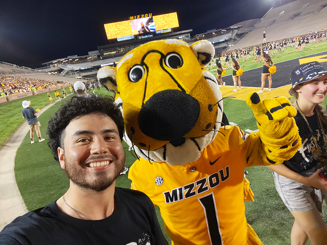 A Hispianic college student wearing black, with his college mascot, Truman the Tiger, on the field at a football game or other event.