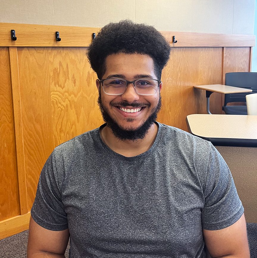 Young African American student with dark hair and a dark beard, wearing glasses and a gray t-shirt, sitting in front of desks.