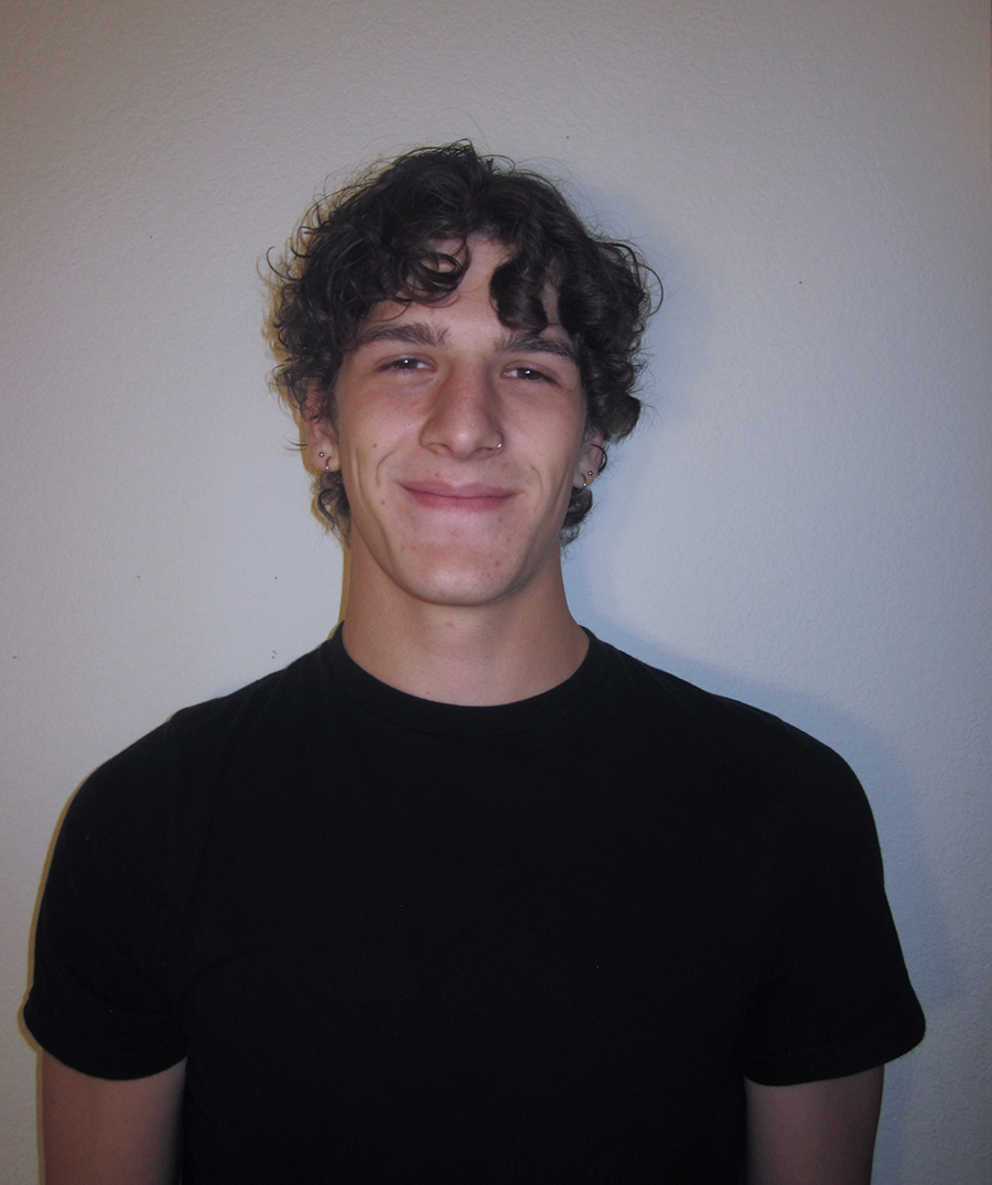 Young male college student standing in front of a wall, smiling. He has dark hair and eyes and curly short hair. He is wearing a black t-shirt.