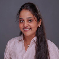 A young Indian college student with long dark and dark eyes. She is smiling. She is wearing a pink button down shirt with a collar. There is a gray background.