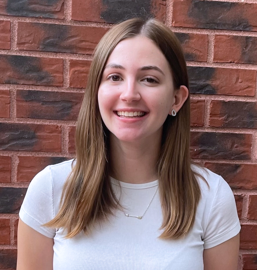 Young college girl with long brownish blonde hair smiling. She is wearing a white tshirt, gold earrings and necklace and is in front of a brick wall.