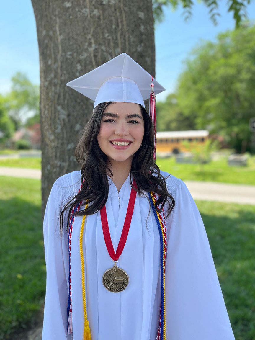 High school student wearing white graduation cap and gown with cords and a medal with red strap. She has brown long hair and is smiling. She is standing in front of a tree.