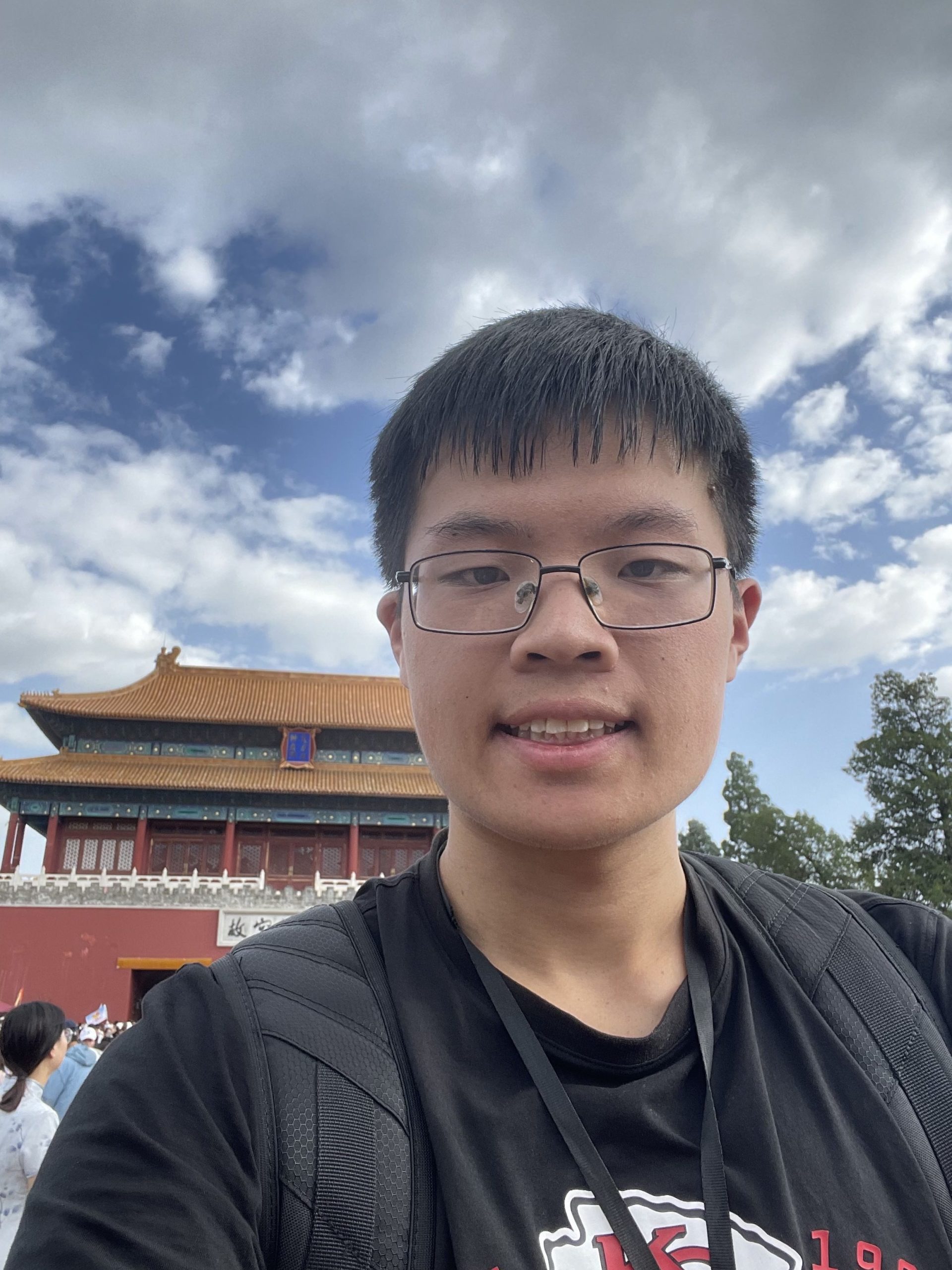 Young Asian man in front of an Asian style building with a bright blue sky with clouds. He is smiling. He wears glasses and has dark hair which is cut to the middle of his forehead. He is wearing a black t-shirt.