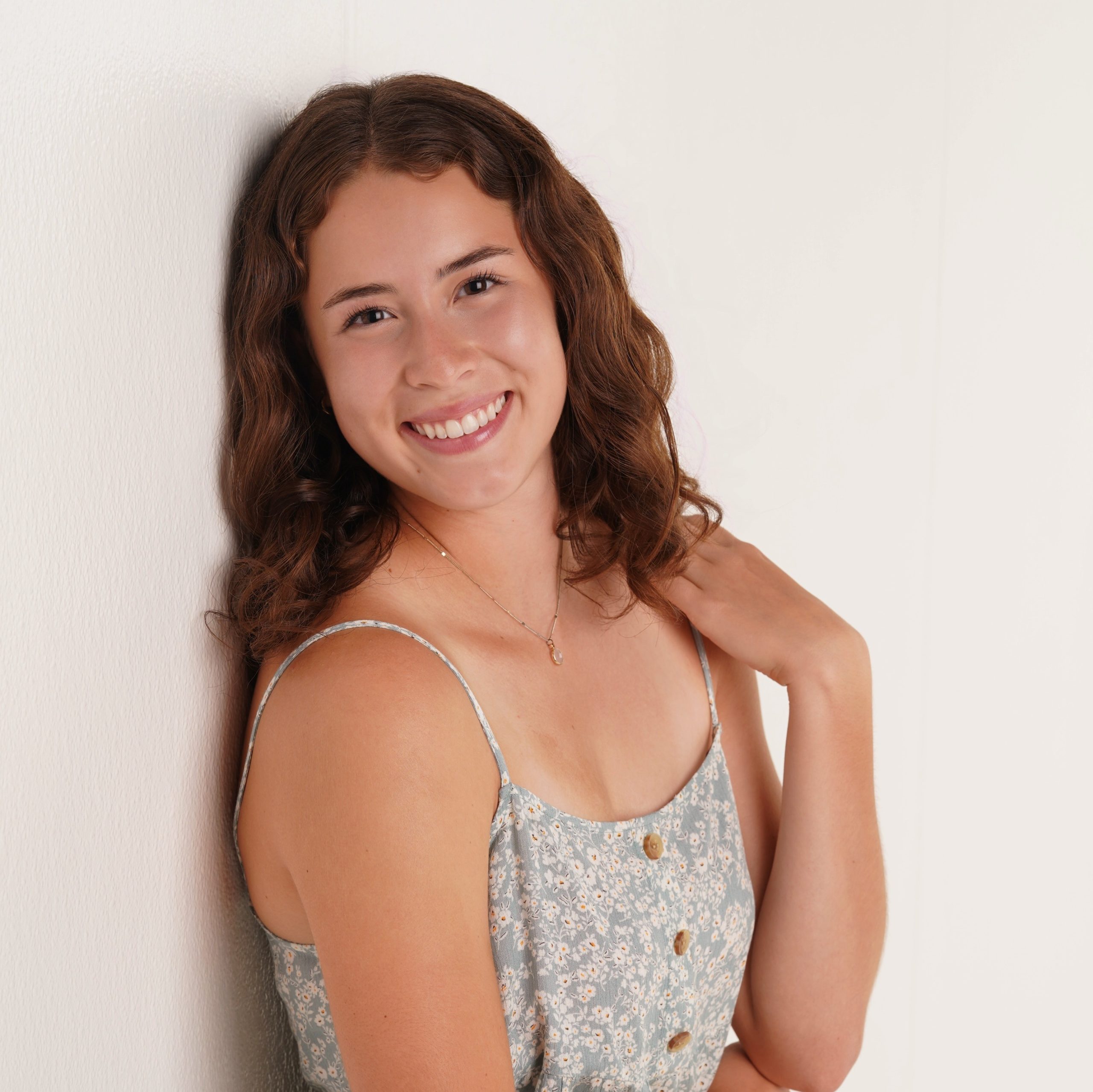 Young lady leaning against a wall. She is a tanned Caucasian, and has her arm folded up and hand placed on her left shoulder. She has long curly brown hair and is smiling.